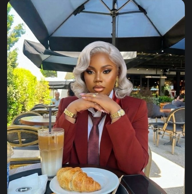 A stylish woman with silver hair, wearing a red suit and tie, sitting at an outdoor café table with an iced coffee, croissant, and phone in front of her.