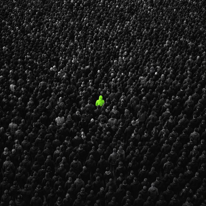 A vast, monochromatic crowd of people stretches across the frame, with a single figure highlighted in vivid neon green standing out in isolation at the center. The image visually echoes the post's theme of "Loneliness," emphasizing one individual dwarfed and set apart by the sea of darkness around them.