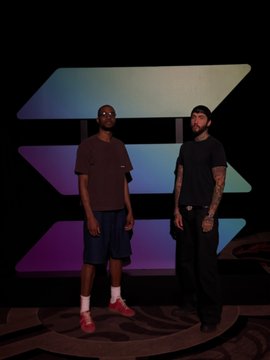 Two men stand side by side in a dimly lit space in front of an illuminated, angular sign featuring a stylized "S" logo—the Solana blockchain emblem—with a colorful gradient backdrop. The post context identifies the pair as the Solana CTO and a colleague, posing for what appears to be a promotional or interview photo.