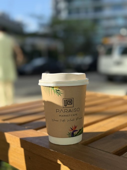 A takeaway coffee cup from 'PARAISO MARKET CAFE' sitting on a wooden table outdoors with a blurred street scene in the background.