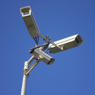 The image features a street lamp pole with three cameras attached to it, all facing upwards towards the sky. The cameras are positioned at different heights and angles, suggesting that they are designed for surveillance purposes. In addition to the cameras, there is also a small text in the image that reads "Crypto(dot)com partners with Trillium Surveyor to expand surveillance and manipulation monitoring across prediction markets". This could be an advertisement or promotional message related to the street lamp pole's surveillance capabilities.