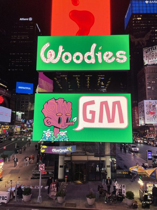A nighttime view of Times Square billboards featuring a large green "Goodies GM" advertisement with a pink cartoon character and flower, alongside other ads including an Apple logo and Broadway show promotions.
