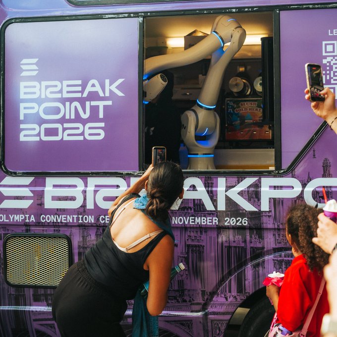 A woman takes a photo of a robotic arm inside a purple 'BREAKPOINT 2026' promotional vehicle at an event scheduled for November 2026.