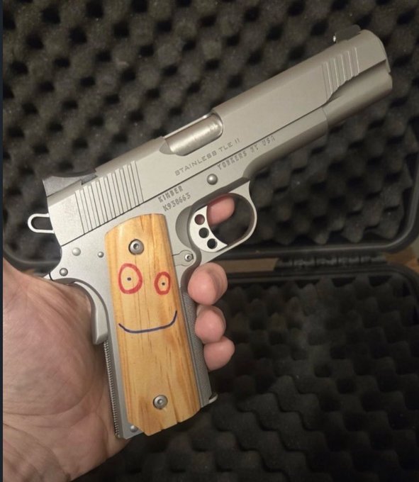 A hand holds a stainless steel semi-automatic pistol marked "STAINLESS TLE II" and "TALGER 37"94" with serial number K959865, featuring wooden grip panels decorated with a hand-drawn red and black smiley face.