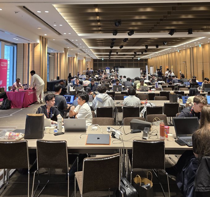 A conference or hackathon event in a large hall, with many attendees seated at long tables working on laptops and networking.