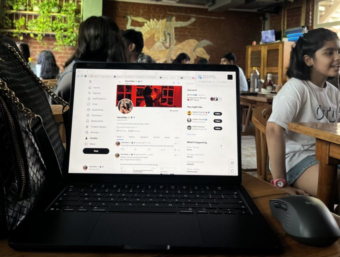 A laptop on a wooden table displays what appears to be an X (formerly Twitter) profile page, with the setting resembling a cozy café or restaurant featuring a brick wall with a painted mural in the background. A young girl sits nearby, smiling and engaged in conversation, embodying the post's theme of "The Art" (the digital content displayed) and "The Artist" (the person creating or sharing it).