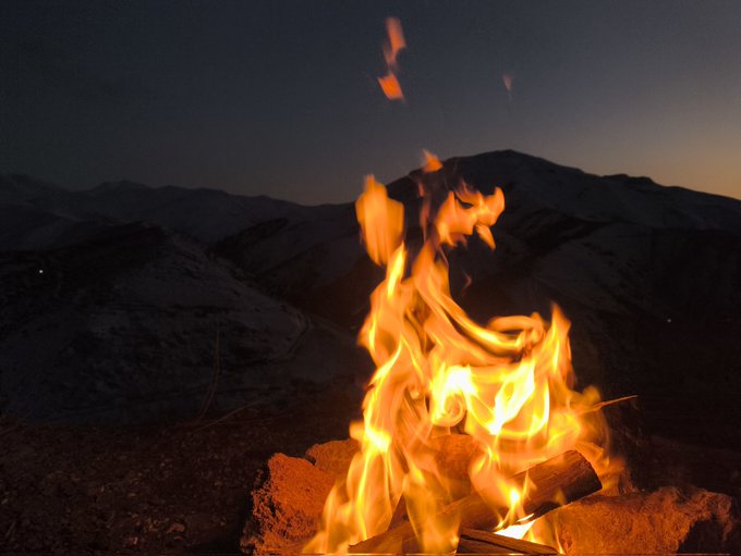 The image shows an open fire burning brightly with a mountain range in the background. The fire appears to be located near a campsite or outdoor area where people are enjoying their time, possibly watching a movie or engaging in other activities during the night. The scene suggests that the campers have gathered around the fire for warmth and entertainment as they spend quality time together outdoors.