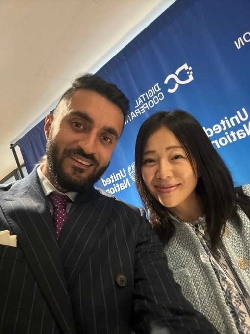 Selfie of a man in a dark pinstripe suit with a purple tie and a woman in a patterned blouse, posing in front of a blue 'United Nations Digital Cooperation' backdrop.