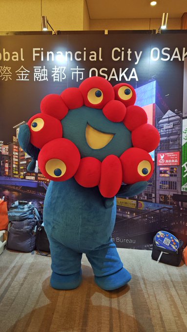 A blue mascot character with a large red flower-like head featuring multiple eyes, posing in front of a banner that reads "Global Financial City OSAKA" and "国際金融都市OSAKA."