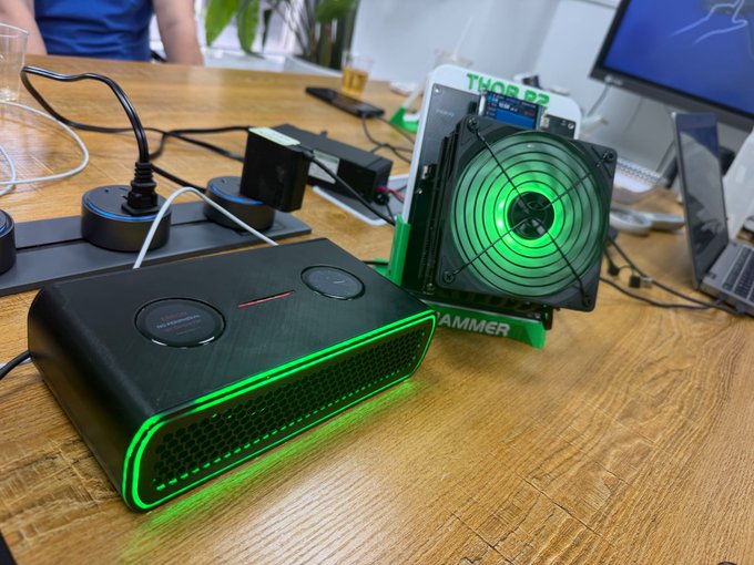 A gaming/PC setup on a wooden desk features a black 'HAMMER' branded device with green LED accents and a cooling fan with matching green illumination.