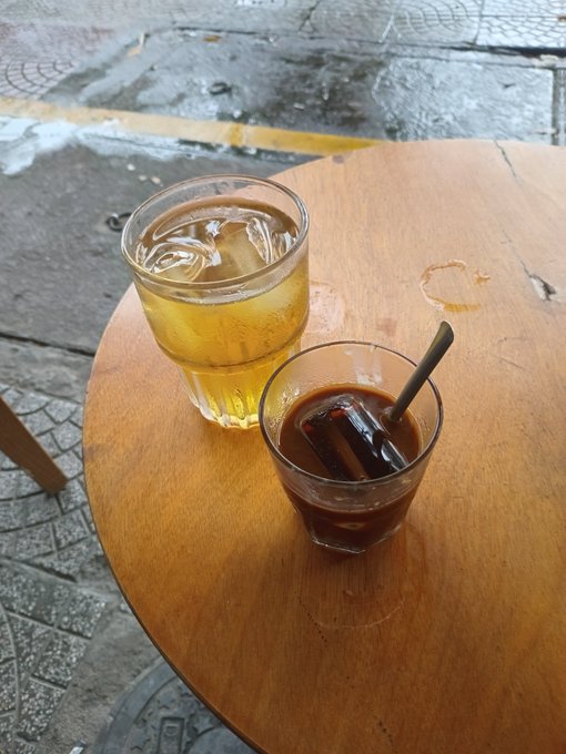 Two glasses of iced beverages—one light-colored tea and one dark iced coffee with a straw—sitting on a round wooden table on a sidewalk.