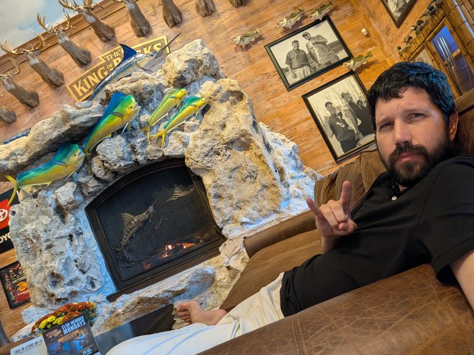 A man sits in a brown leather chair making a hand gesture in front of a stone fireplace decorated with colorful fish mount sculptures and a 'King of Beers' sign, with framed photos on the wooden wall behind him.