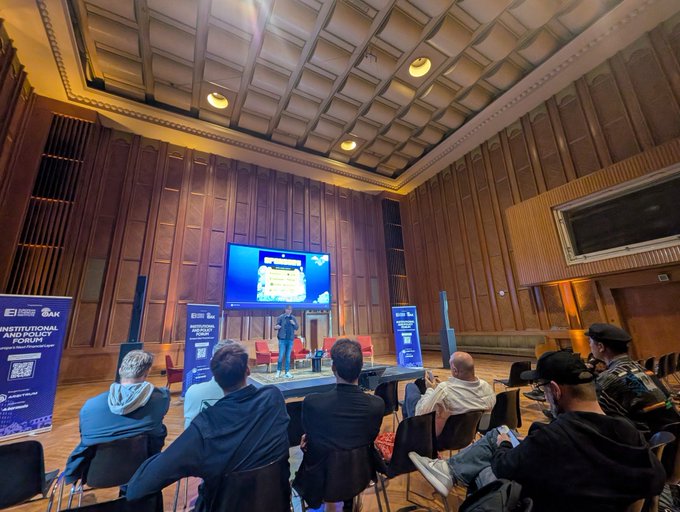 The image shows a group of people sitting in chairs, watching a presentation or event taking place on a large screen TV mounted high up on the wall. The room has wooden walls and ceiling, giving it an old-world charm. There are several people seated around the room, some standing near the back while others sit closer to the front. A few individuals can be seen holding handbags as they enjoy the event or presentation. The atmosphere appears to be relaxed yet engaged, with everyone focused on what is being displayed on the screen.