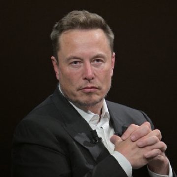 A portrait of Elon Musk wearing a black suit jacket over a white shirt, with his hands clasped in front of him against a dark background.