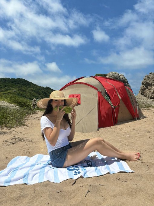 A woman in a sun hat and white top sits on a striped beach towel holding a slice of watermelon, with a red dome tent pitched on the sandy shore behind her under a partly cloudy blue sky—capturing a relaxing beachside camping moment perfect for a cheerful "Hump Day" greeting.