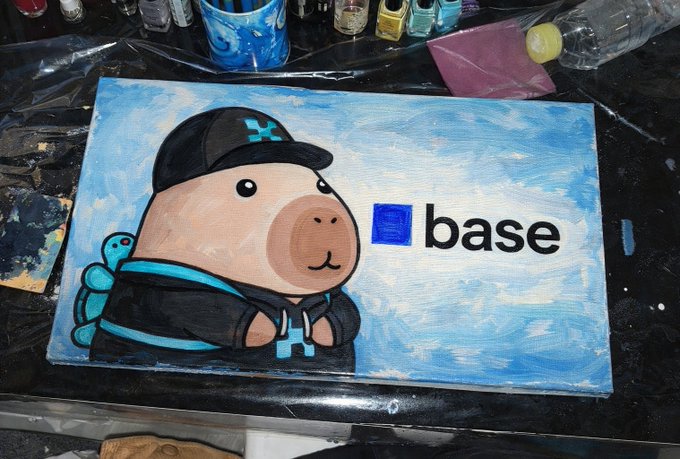 The image features a cartoon drawing of a pig wearing a hat with the word "base" written on it. This drawing is placed on top of a table, surrounded by various art supplies such as paintbrushes, cups, and bottles. The scene suggests that someone has been working on an art project or craft, possibly creating a unique piece inspired by the concept of Base.