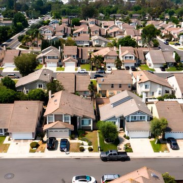 An aerial view of a suburban neighborhood featuring rows of houses with tiled roofs, some equipped with solar panels, along with parked cars and green trees.