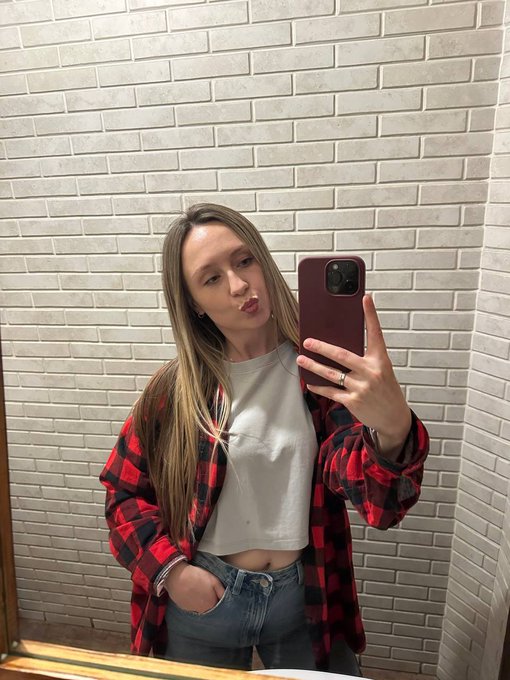 A blonde woman in a red and black plaid flannel over a white t-shirt takes a mirror selfie in a bathroom with white brick tile walls.