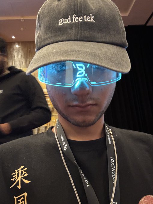 A person wearing a 'gud fee tek' cap, futuristic glowing cyan smart glasses, and a black shirt with Asian characters poses at what appears to be the TOKEN2049 crypto conference.