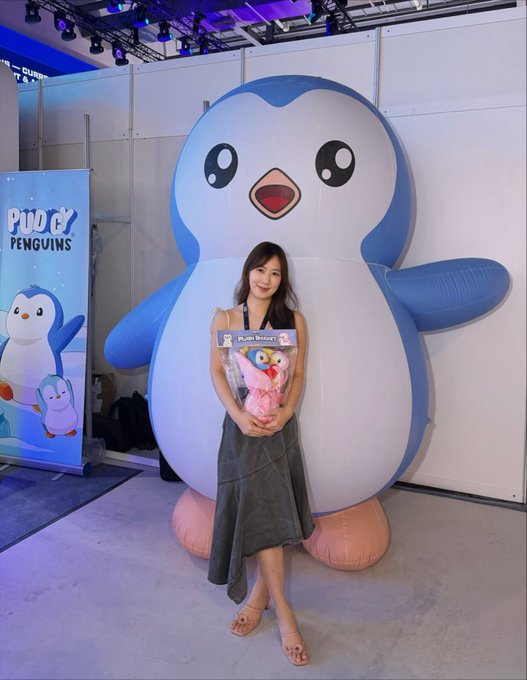 A woman poses in front of a large inflatable Pudgy Penguin mascot at the Pudgy Penguins pop-up booth, holding a plush penguin bouquet that matches the event's branding. The booth's banner displays the "Pudgy Penguins" logo alongside cartoon penguin illustrations, consistent with the Ultra Web3 Festival promotion.