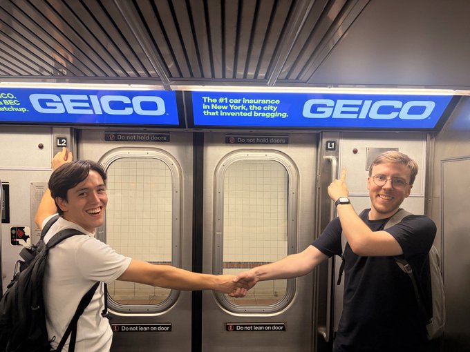 Two smiling men shake hands and point at GEICO advertisements inside a New York City subway car, with one sign reading 'The #1 car insurance in New York, the city that invented bragging.'