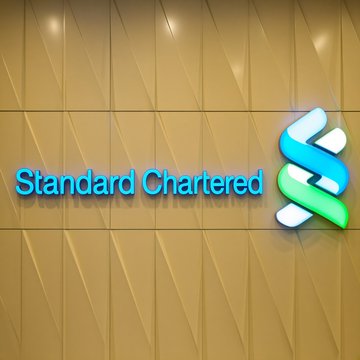 A photograph of the Standard Chartered bank sign mounted on a golden, textured wall, featuring their blue and green intertwined logo.