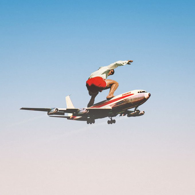 A surreal, humorous photo composite showing a man in red shorts and a white sweatshirt crouched in a surfing pose on top of a commercial 'Smarat' airplane in mid-flight.