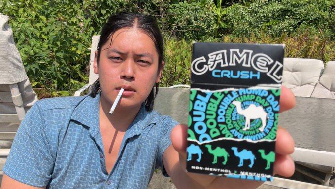 A man smokes a cigarette while holding up a pack of Camel Crush Double Menthol cigarettes, juxtaposing the harmful habit with a satirical "antidote" reference to memecoins as financial cancer in the accompanying video.