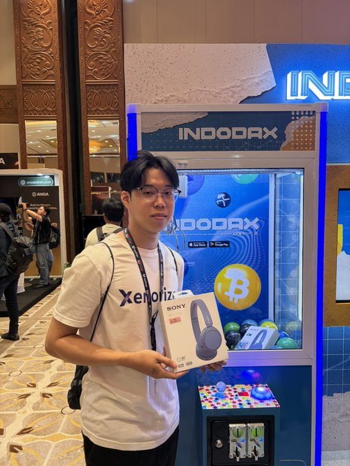 A man wearing glasses and a white t-shirt poses proudly with a Sony CH-520 wireless headphone box in front of the INDODAX booth featuring Bitcoin (₿) branding, celebrating his Day 2 win at the IDBW event by Indodax.