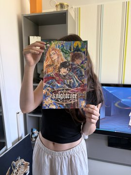 A person holding up a 'Finalcore' art book or manga, covering their face, standing in a room with a computer monitor and shelving visible.