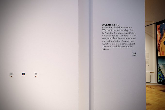 A museum or gallery interior showing a white wall with German text titled 'AGENT NETS' explaining blockchain-based works connecting autonomous digital AI agents, alongside a QR code.