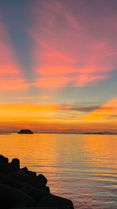 A vibrant sunset over a calm ocean with orange and pink clouds reflected on the water, featuring silhouettes of rocks in the foreground and a small island on the horizon.