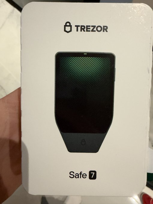 A hand holding a white retail box for a Trezor Safe 7 hardware wallet, displaying an image of the black device with a green geometric screen.