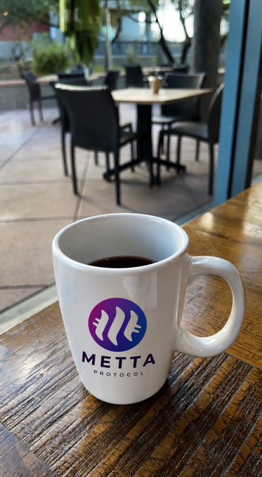 A white coffee mug with a purple 'METTA PROTOCOL' logo sits on a wooden table, with outdoor patio seating visible through a window in the background.