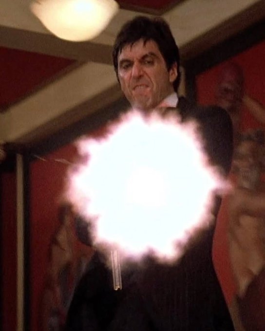 A movie still of a man in a tuxedo grimacing as he shoots a handgun, with a large muzzle flash visible in the foreground.