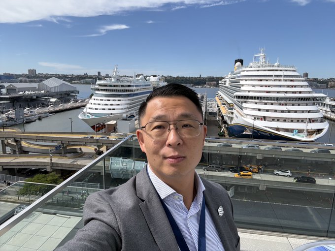 A man in a gray blazer and glasses takes a selfie at a cruise terminal with two large cruise ships docked behind him and a cityscape in the background.