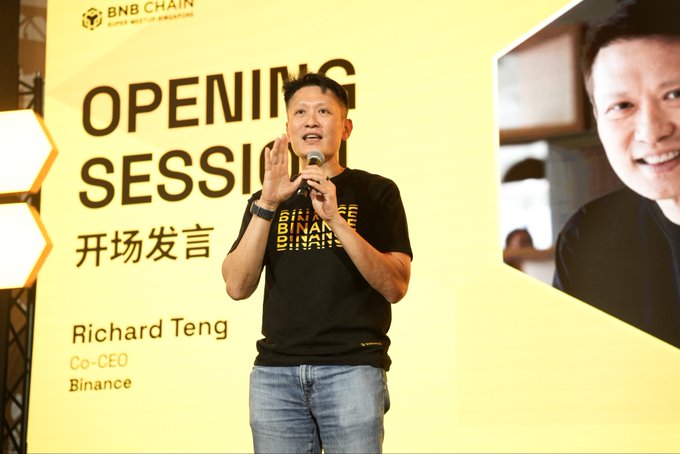 Richard Teng, Co-CEO of Binance, delivers an opening session speech at the BNB Chain #BNBSG event while wearing a black Binance-branded t-shirt. A large screen behind him displays the BNB Chain logo and "OPENING SESSION / 开场发言" text alongside his photo, with a second image showing him smiling.