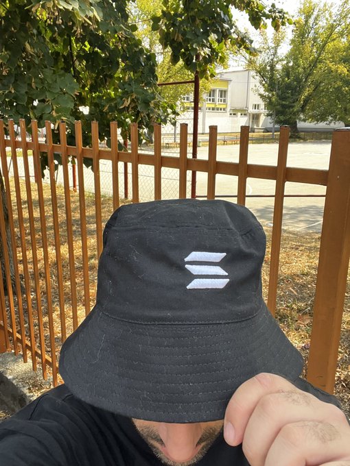 A selfie of a person wearing a black bucket hat featuring the Solana logo, photographed outdoors near a wooden fence with trees in the background.