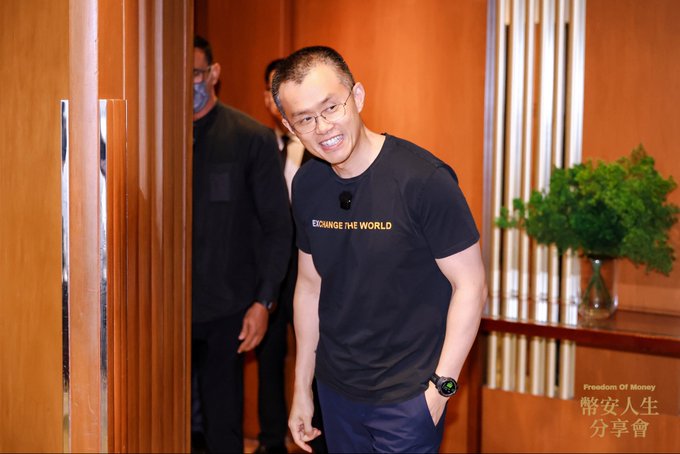 A smiling man wearing glasses and a black t-shirt reading 'EXCHANGE THE WORLD' stands in a doorway, with text in the bottom right corner reading 'Freedom Of Money 幣安人生 分享會'.