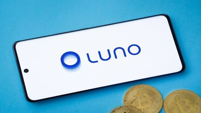 A smartphone displaying the 'Luno' logo rests on a blue surface next to two physical Bitcoin coins.