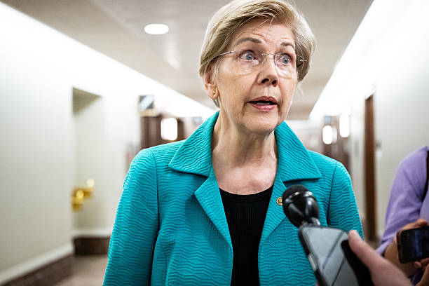 Senator Elizabeth Warren, wearing glasses and a teal blazer, speaks to reporters in a hallway with microphones held up to her. The image connects to the post's humorous jab at her skepticism toward cryptocurrency, suggesting she is unaware that Bitcoin's open-source code is publicly available rather than controlled by a single corporate entity.