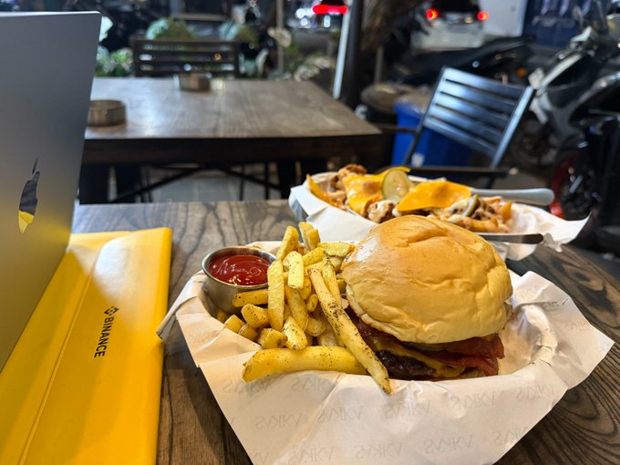 A casual restaurant scene shows a wooden table with a burger and seasoned fries in a paper-lined basket next to ketchup, alongside a yellow Binance-branded paper bag, with a laptop and motorcycle visible in the background.