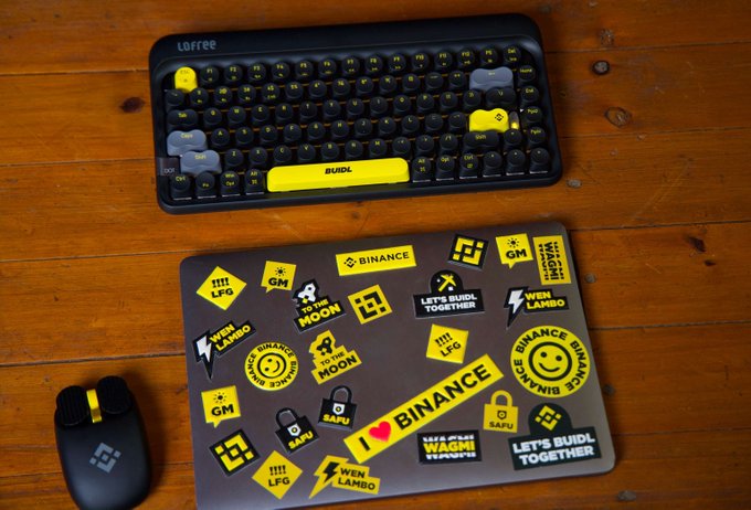 A mechanical keyboard with yellow accent keys and the word 'BUIDL' sits above a mousepad covered in cryptocurrency-themed stickers, including Binance, SAFU, and 'WAGMI' slogans.