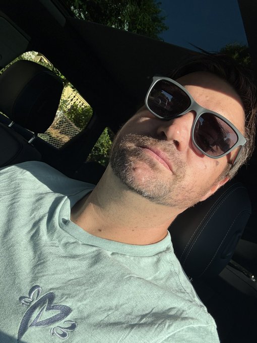 A man wearing sunglasses and a light green t-shirt featuring the MetaVixens logo relaxes in the driver's seat of his car, showing off the new NFT merchandise in the sunshine.