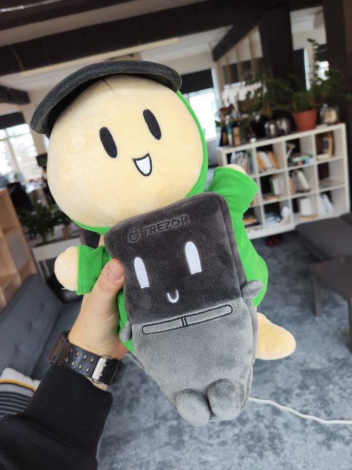 A hand holds up two plush toys in a modern living room setting: a green character and a smaller, grey Trezor hardware wallet mascot with a smiling face.