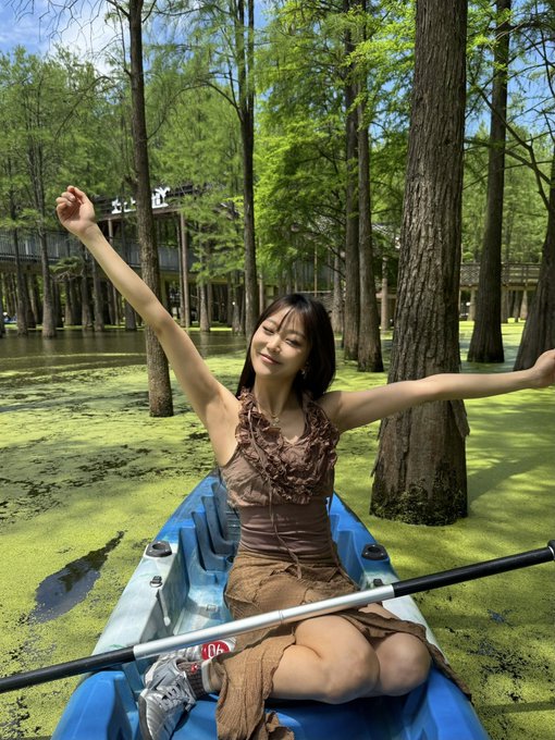 A young woman in a brown ruffled dress joyfully poses with arms outstretched while kayaking through a lush, green swamp filled with tall trees.