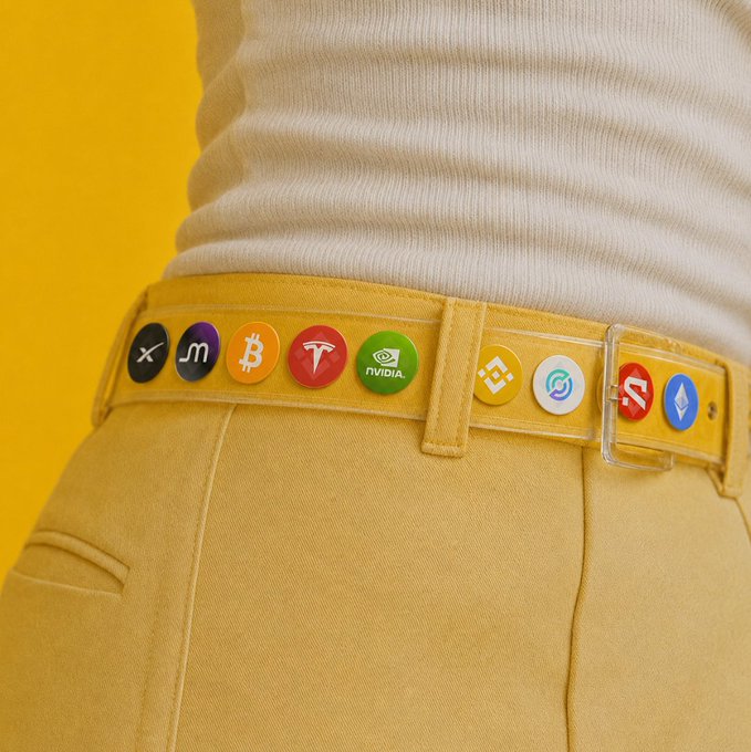 The image features a person wearing yellow pants, with a yellow belt adorned with various colored buttons including red, green, blue, and white. The person's shirt has logos of Bitcoin, Tesla, and Nintendo on it. The background appears to be a yellow wall or surface.