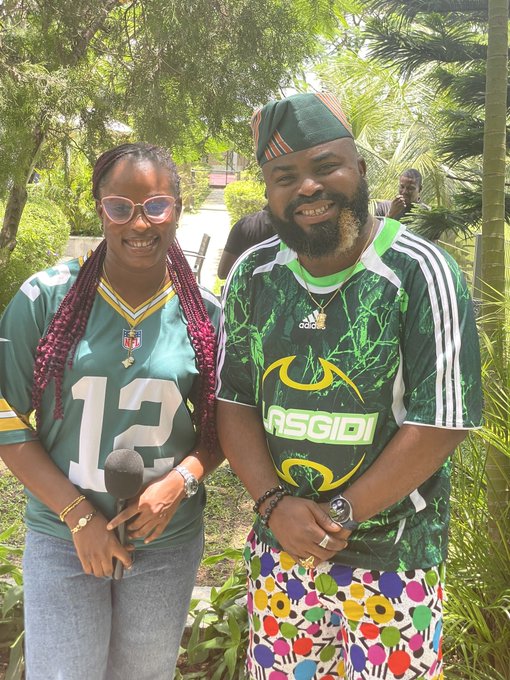 Two people smiling outdoors in a tropical garden; the woman wears a Green Bay Packers #12 jersey with heart-shaped sunglasses and holds a microphone, while the man wears a green Nigerian-style Adidas jersey and patterned shorts.