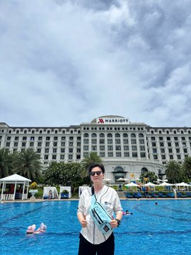 A person stands in front of a large Marriott hotel with a swimming pool in the foreground, holding a package.