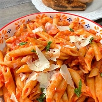 Close-up of a plate of penne pasta in a pink tomato-cream sauce, topped with shaved parmesan cheese and fresh herbs, with bread visible in the background.